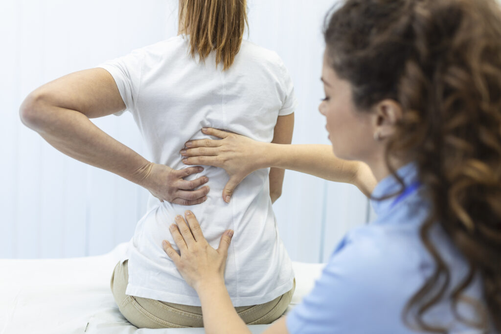 Profissional de saúde realiza avaliação da coluna de uma paciente durante consulta para diagnóstico e indicação de cirurgia minimamente invasiva.